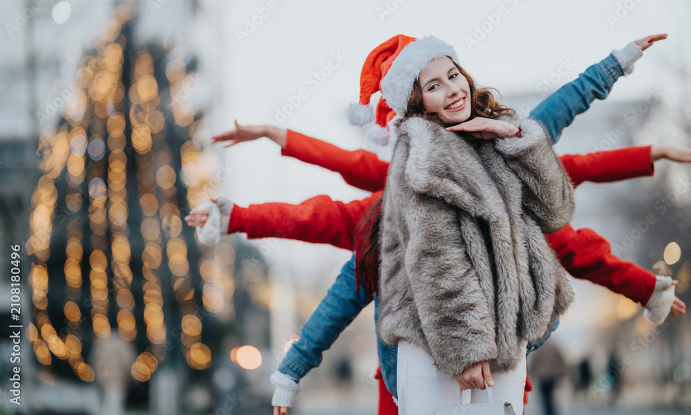 Fototapeta premium Three girls wearing Santa hats pose together outdoors with joyful expressions. The friends celebrate a festive holiday evening with blurred lights in the background.