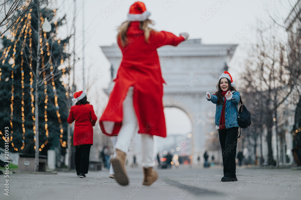 Fototapeta premium Two girls greet each other with open arms wearing Santa hats and winter coats. A festive city street scene with holiday lights and cheerful expressions.