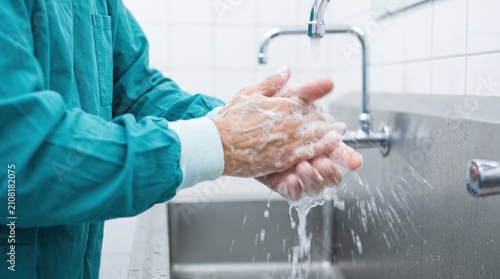 Surgeon washing hands with soap and water under a faucet in a hospital scrub room before a medical operation, representing healthcare hygiene and sterilization