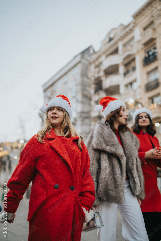 Fototapeta premium Three girls wearing Santa hats and winter coats walk along a city street. A group of friends in festive outfits stroll past urban buildings for the holidays.