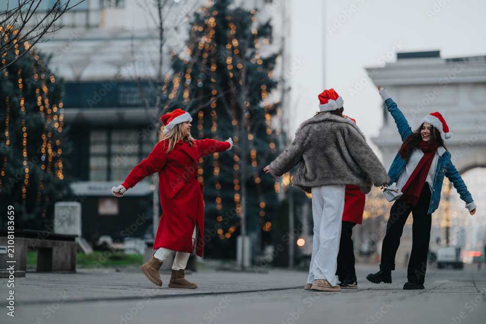 Fototapeta premium Three girls wearing Santa hats laugh and dance together on a city street decorated with lights. The festive friends celebrate a winter evening under holiday lights and archways.