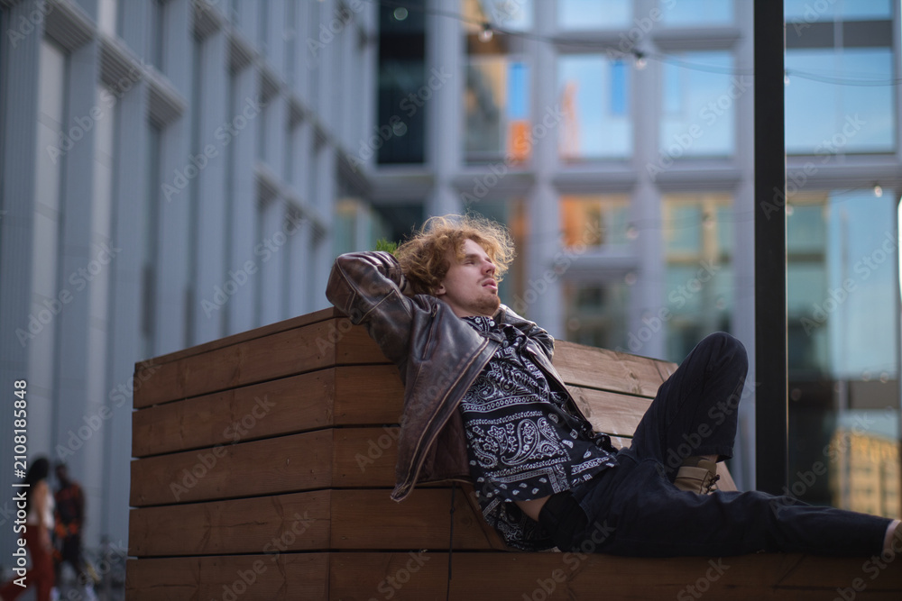 Fototapeta premium Relaxed young man in a leather jacket sitting on a wooden bench in a modern urban environment. Authentic lifestyle portrait representing freedom, confidence, relaxation, contemporary city life
