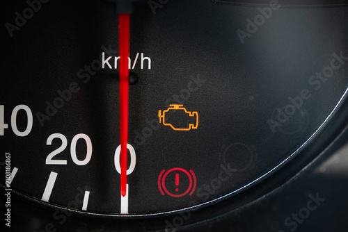 Close up view of an illuminated check engine light and hand break on warning signal on a black car dashboard speedometer scale.