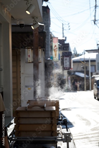 Photography 湯気の立つ木製蒸し器と路地の生活感を捉えた地方の街並み  
Steaming wooden food boxes along a sunlit local al