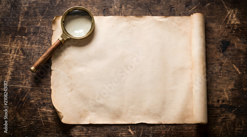 Blank vintage parchment paper scroll with an antique magnifying glass on a dark rustic wooden table background with copy space