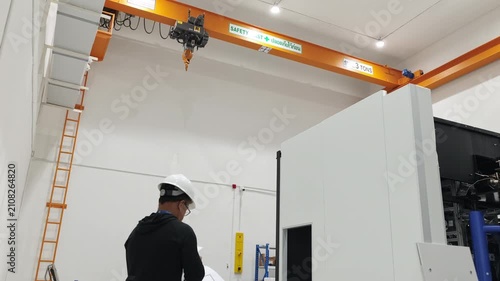 An engineer wearing a white hardhat and safety glasses reviewing documents inside a modern industrial facility equipped with a yellow overhead traveling crane