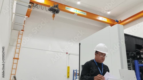 An engineer wearing a white hardhat and safety glasses reviewing documents inside a modern industrial facility equipped with a yellow overhead traveling crane