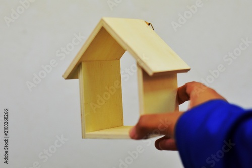 A hand holds a small, hollow wooden house model against a neutral background, symbolizing real estate and housing concepts.