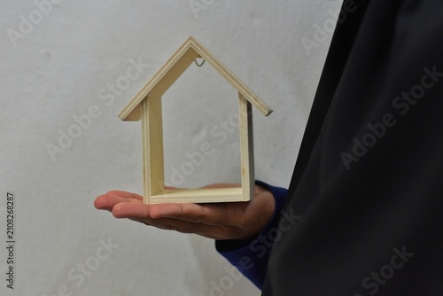 A person holds a small, open-frame wooden house model in their hand against a plain background, representing real estate.