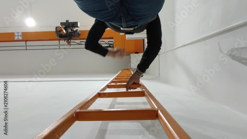 A low-angle view looking up at a person in jeans and a black shirt climbing an orange metal ladder in an industrial facility, moving upward toward an overhead crane