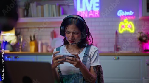 Asian young e-sport woman gamer play online mobile game on smartphone.Attractive girl vlogger influencer content creator gamer enjoy and fun live streaming broadcast games in POV screen and neon room.