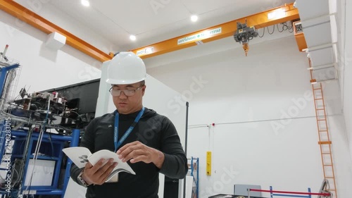 An engineer wearing a white hardhat and safety glasses reviewing documents inside a modern industrial facility equipped with a yellow overhead traveling crane