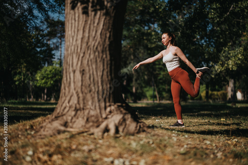 Konstfotografi Woman performing a standing quad stretch in a sunny park to warm up for exercise