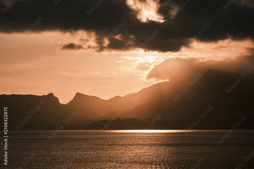 Fototapeta premium Breathtaking sunset over a tranquil lake, framed by majestic mountains and dramatic clouds. Golden light beams pierce through, creating a serene and powerful natural landscape.