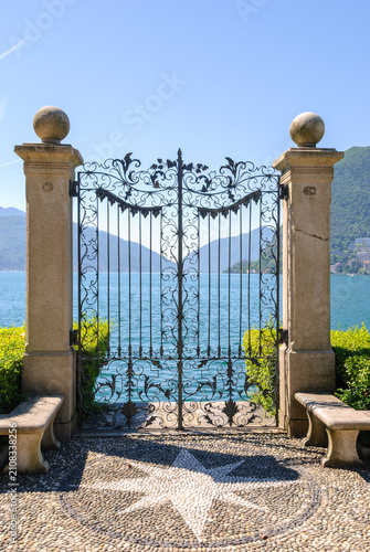 Elegant wrought iron gate overlooking Lake Lugano in Lugano, Switzerland, framed by mountains and blue sky, showcasing timeless architecture and serene lakeside scenery.