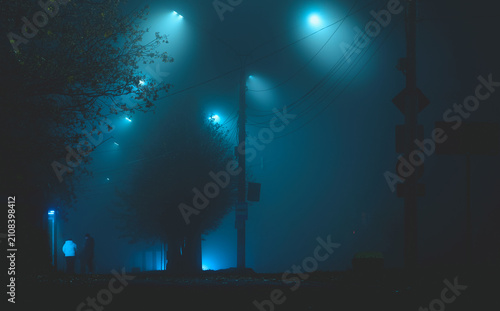 Cinematic night street in thick fog with silhouettes of people and glowing cyan streetlights cutting through the mist, moody urban background.
