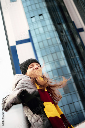 Obraz na płótnie Low angle portrait of a smiling young woman with long windblown hair, wearing a striped Gryffindor style scarf, beanie, and winter jacket against a modern glass skyscraper
