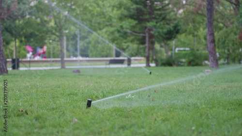 Sprinkler system watering green grass in a park, with visible trees and blurred figures enjoying leisure activities in the background during a sunny day