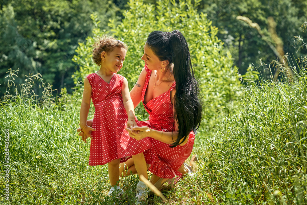 Fototapeta premium A mother and daughter wearing matching red-polka-dot dresses are walking in a summer park. The girl is laughing, her eyes shining with joy and curiosity. Sunlight filters through the tall green grass.
