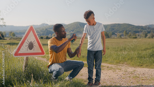 A man sprays tick repellent on his sons arm during an outdoor activity in a grassy field preventing tick bite.