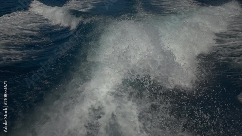 Turbulent white foam water trail behind a large boat in a calm blue sea on a sunny day, slow motion