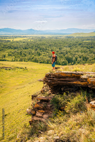 Hiker with backpack stands on edge of mossy cliff, admiring endless panorama of forest-steppe with mountains on horizon. A solo traveler without face, looking into distance, slow travel