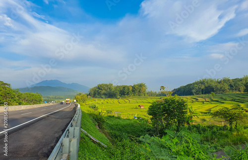 Scenic toll road beside lush green rice fields in Central Java, Indonesia, under a dramatic sky. Ideal for travel, infrastructure, and distribution.