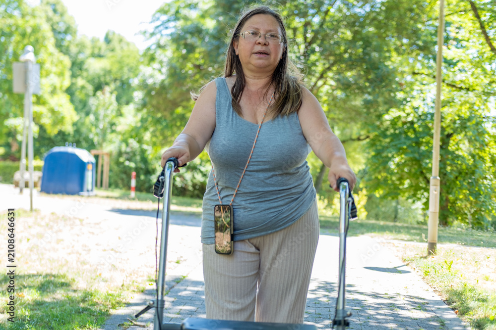 Obraz premium Middle-aged woman walking rollator paved path, mobility aid assistance and independence, Rehabilitation therapy exercises outdoors, activity disabled person moving independently, physical limitations
