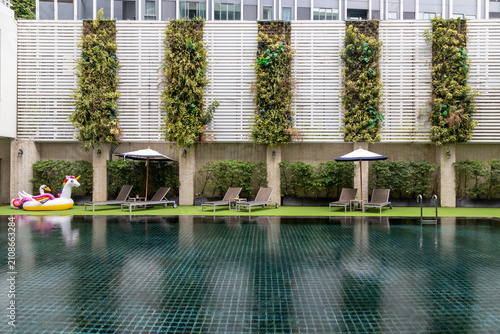 A scenic swimming pool area featuring dark tiled water, multiple sun loungers, umbrellas, a unicorn inflatable float, artificial turf, and a striking vertical garden wall with white slatted panels.