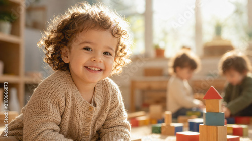 Toddler with curly hair smiles while playing with wooden building blocks in a brightly lit preschool classroom setting.