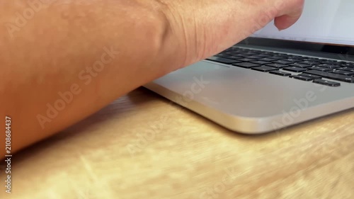 A woman's hand typing text on the keyboard of a laptop standing on a wooden table against a blurred plant. Concept: Remote work, office tasks, typing, business activity. High quality 4k footage