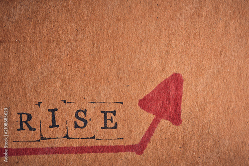 Stamped word RISE with bold red arrow pointing up on textured brown cardboard, macro shot.