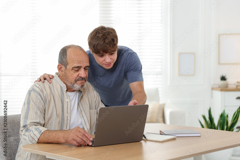 Fototapeta premium Caregiver. Teenager helping senior man learn how to use laptop indoors, space for text