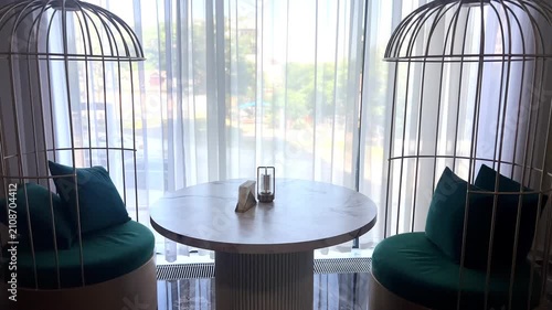The camera slides smoothly to the right, revealing an elegant interior with a round table and unique cage chairs with turquoise cushions against the backdrop of a bright window in a cozy cafe. High