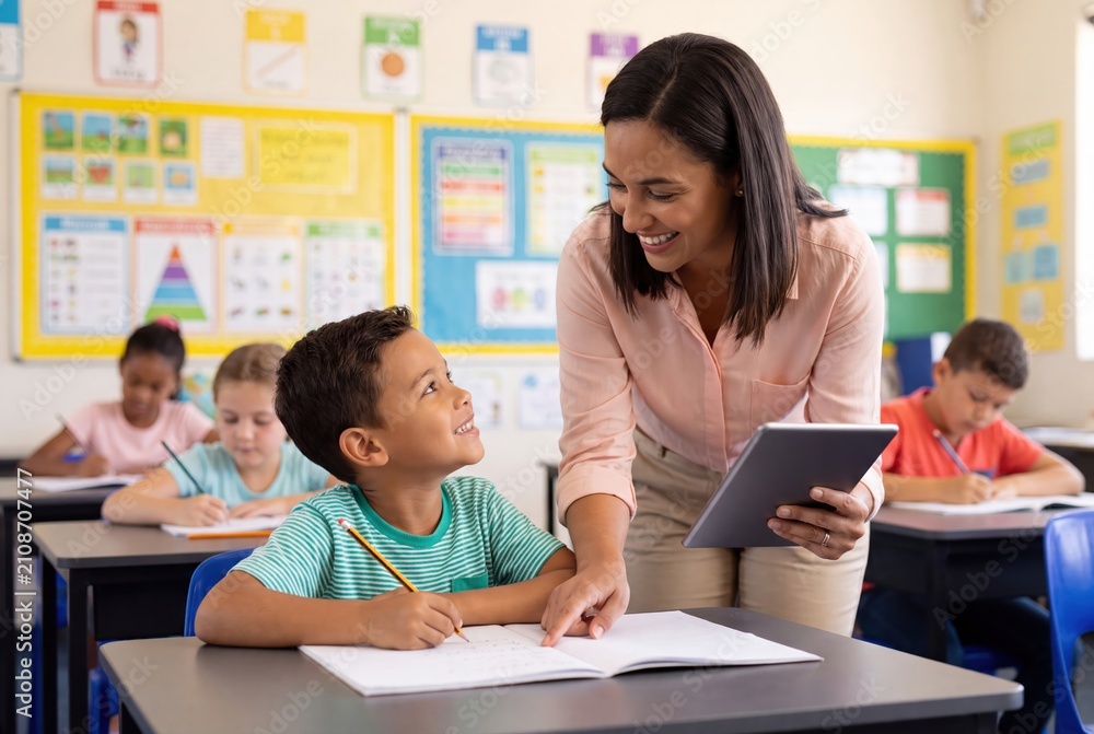 Fototapeta premium A smiling teacher assists a young student with his schoolwork, using a tablet in a bright classroom.