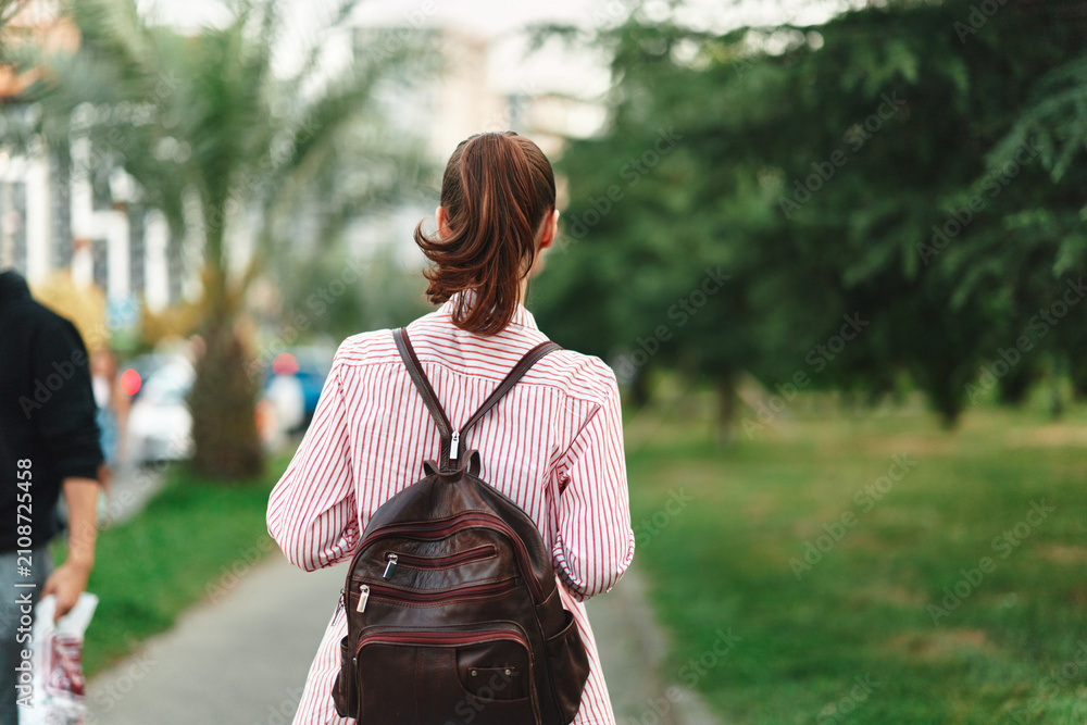 Fototapeta premium Young woman with a backpack walks through a green park from behind, wearing a striped shirt, capturing a casual solo moment of everyday exploration.