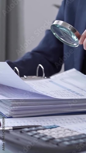 A businesswoman is using a calculator and a magnifying glass to analyze financial documents at her desk in a modern and fair office setting, focusing on aspects of audit and taxes in business