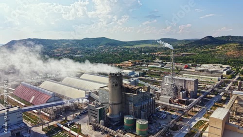 Workers oversee operations at a factory surrounded by large structures and equipment. Smoke rises from chimneys while machinery runs on a clear day.