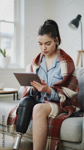 Young woman with prosthetic leg using tablet on sofa with red plaid blanket, medium shot