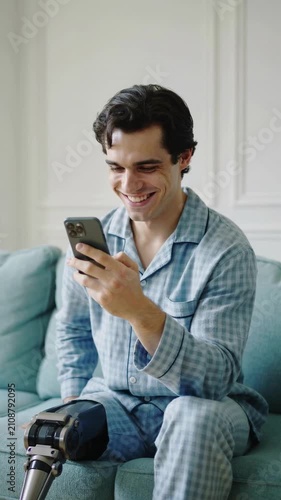 Young man with prosthetic leg sitting on blue sofa using smartphone in interior, medium shot
