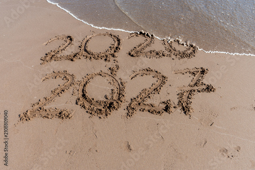 The wave washes away the year 2026 and leaves 2027 written in the sand on the beach.