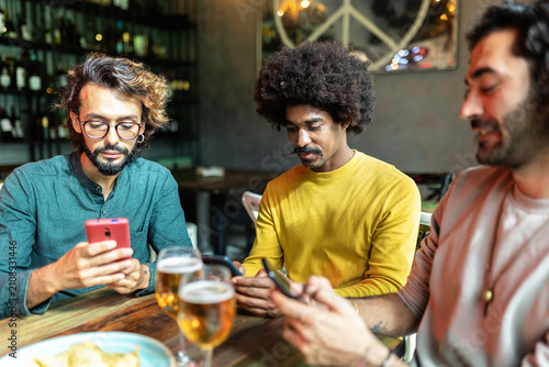 Three male friends using smartphones while drinking beer at a bar, enjoying their evening out together