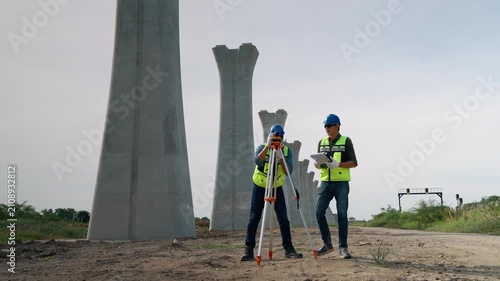 Survey engineer using surveying equipment on a tripod while another engineer reviews project documents beside large concrete bridge columns at a transportation infrastructure project.
