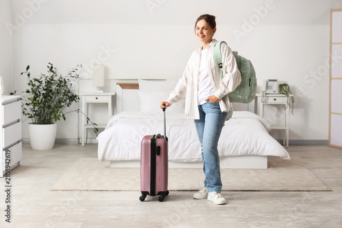 Young woman traveler with a pink suitcase and backpack standing in a bright hotel room, ready for departure