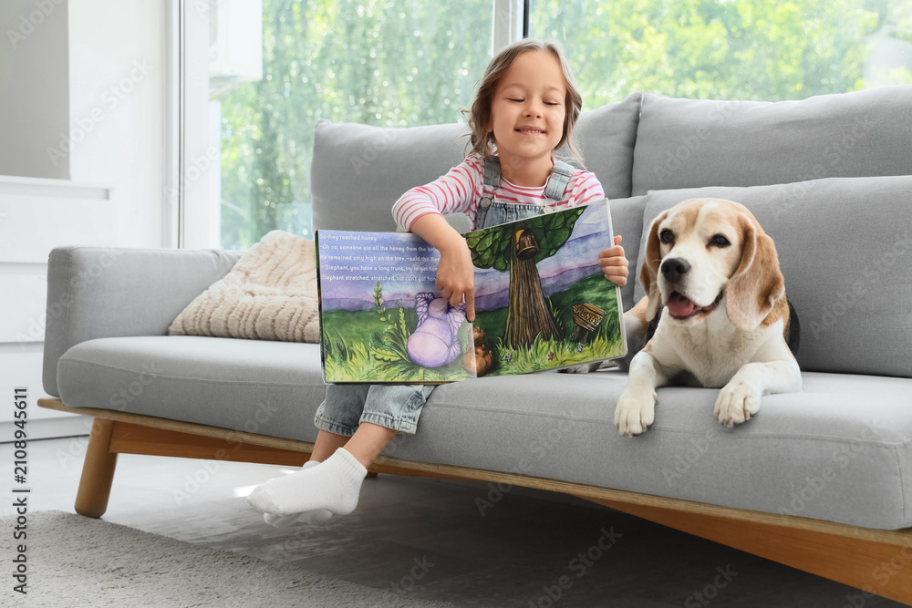 Obraz premium Cute little girl with Beagle dog and book on sofa in living room
