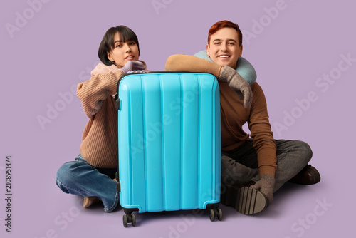 Beautiful young couple with suitcase sitting on purple background. Winter vac...