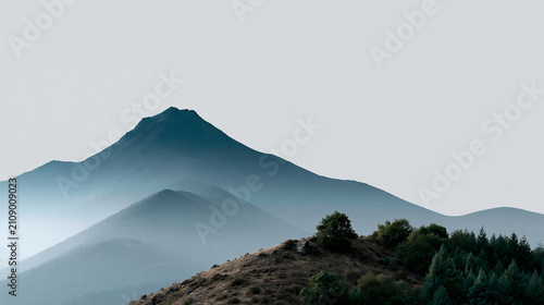 Mountain peak in mist above forest hill, soft blue landscape with serene sky