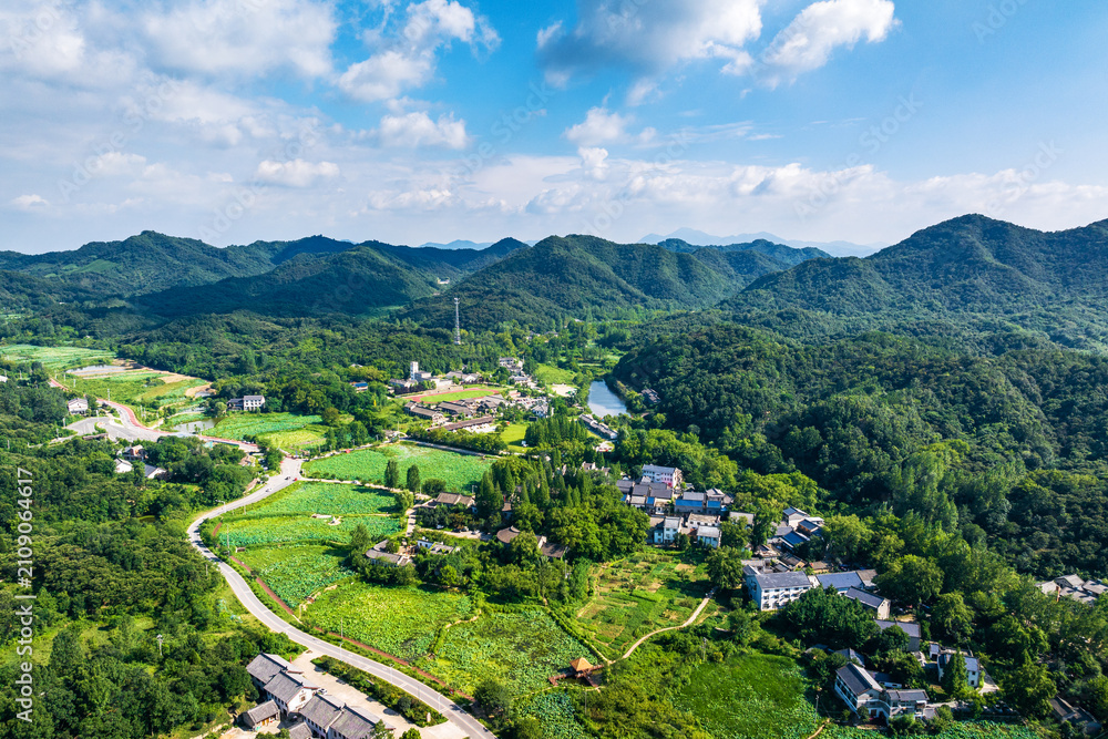 Obraz premium An aerial shot of a rural village nestled among lush green mountains, farmlands and forests under a bright blue sky with white clouds.