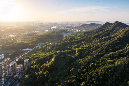 Obraz na płótnie An aerial panorama captures lush green mountains adjacent to urban buildings, illuminated by soft sunrise light, with distant cityscapes on the horizon