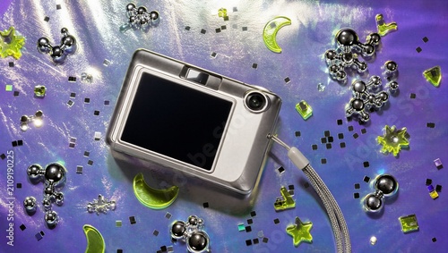 Top view of silver compact digital camera surrounded by chrome atom charms, yellow plastic stars, and confetti on holographic purple background. Y2K nostalgia and retro technology concept